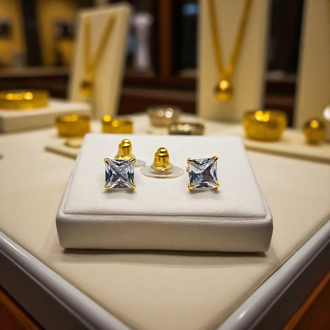 Gold square-cut gemstone earrings for women on a white display box with blurred jewelry store background.