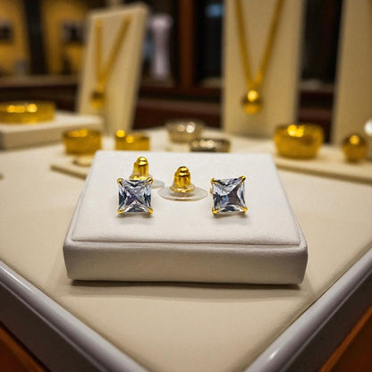 Gold square-cut gemstone earrings for women on a white display box with blurred jewelry store background.