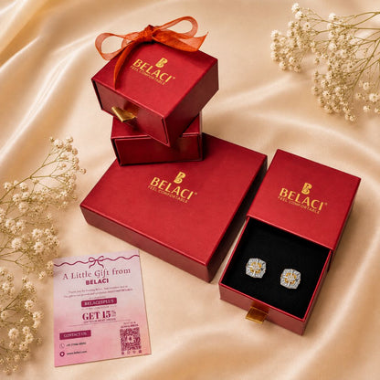 BELACI brand red gift boxes arranged on a satin fabric background, with one open box displaying a pair of square stud earrings in a black insert. A pink promotional card is placed nearby, and dried white flowers decorate the scene.