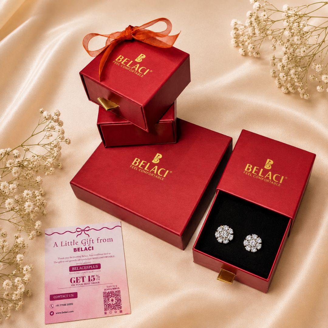 Three red BELACI gift boxes arranged on a satin fabric backdrop, with one open box displaying a pair of round stud earrings on a black insert. A pink gift card is placed in front, and small white floral sprigs decorate the scene.