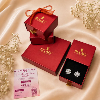 Three red BELACI gift boxes arranged on a satin fabric backdrop, with one open box displaying a pair of round stud earrings on a black insert. A pink gift card is placed in front, and small white floral sprigs decorate the scene.