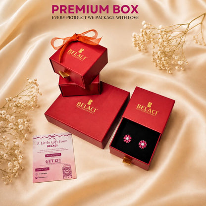BELACI premium jewelry gift box set on draped champagne satin fabric, featuring red boxes with gold branding, one open box displaying pink gemstone earrings, a pink gift card, and decorative baby's breath flowers.