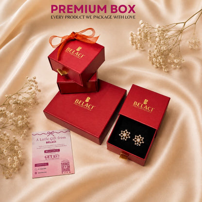 BELACI jewelry gift packaging set on draped champagne satin fabric, featuring red boxes, one open box displaying snowflake-shaped earrings on a black insert, a pink gift card, and dried baby's breath flowers.