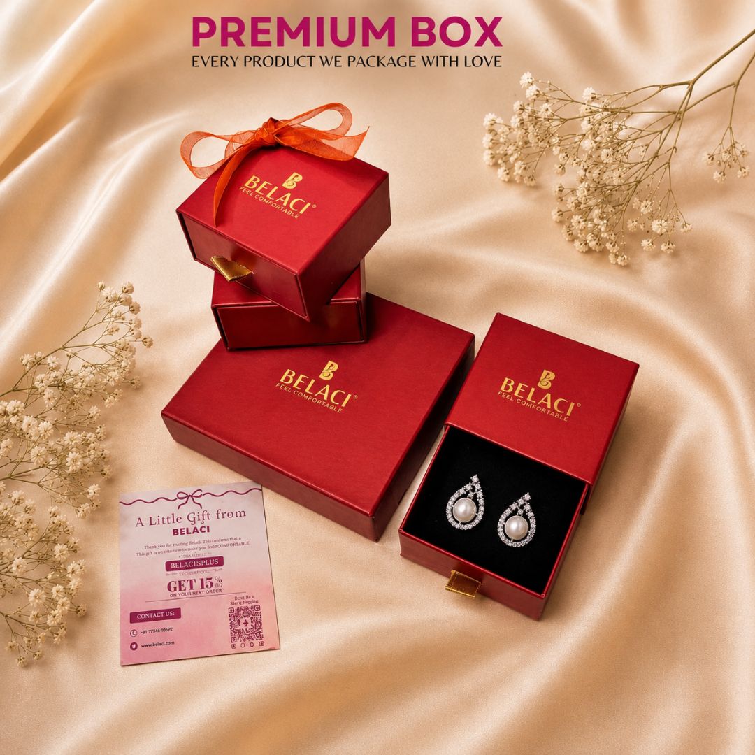 BELACI brand red jewelry packaging boxes, including one open box displaying a pair of teardrop-shaped silver and white gemstone earrings, arranged on a beige satin fabric background with decorative baby's breath flowers and a promotional card.