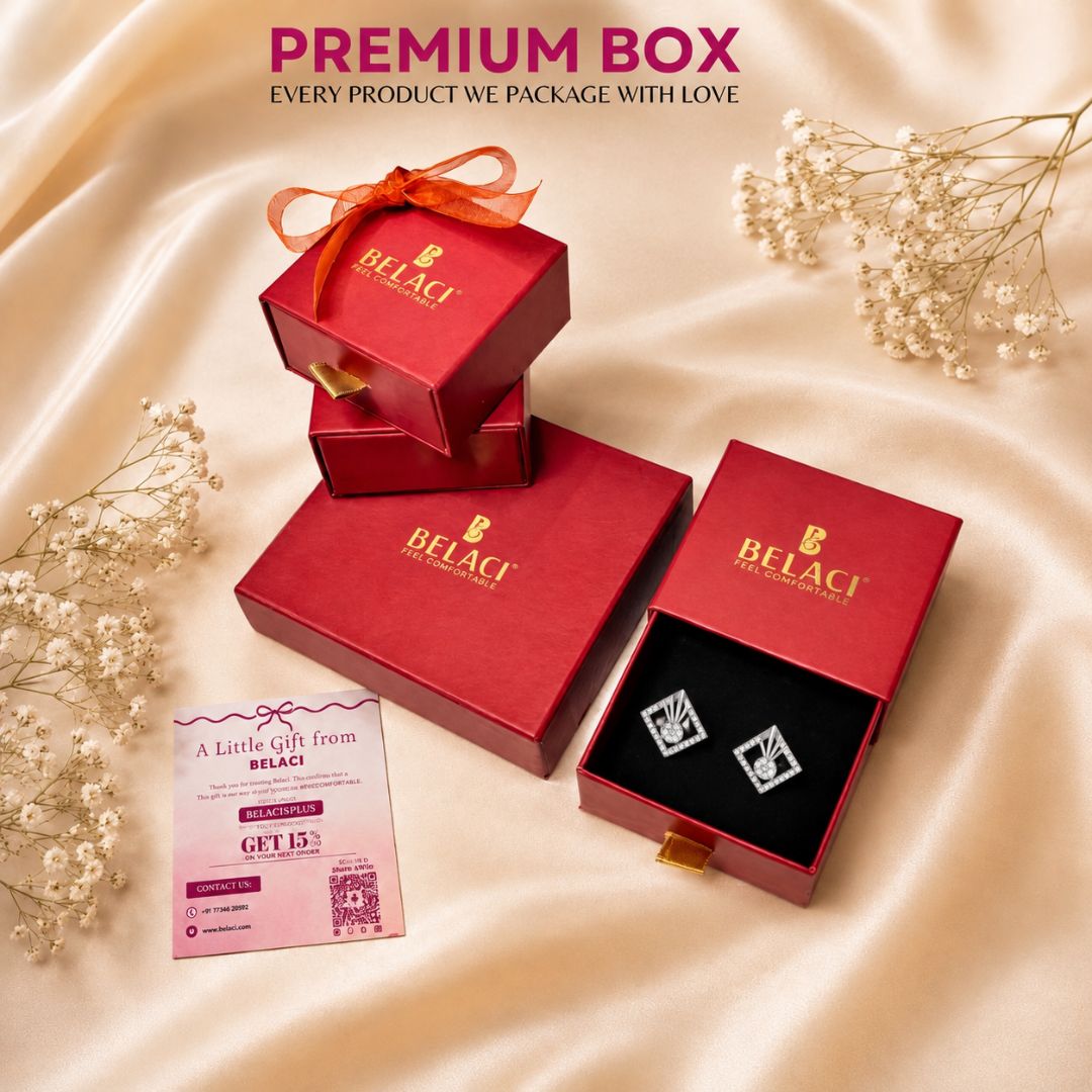 A luxury gift set from BELACI featuring red packaging boxes, one open to reveal silver square stud earrings on a black insert, a gift card, and decorative dried flowers on a beige satin background.