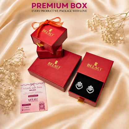 BELACI jewelry gift packaging set on draped champagne satin fabric, featuring red boxes with gold logo, one open box displaying silver floral stud earrings on a black insert, a pink gift card, and dried baby's breath flowers.