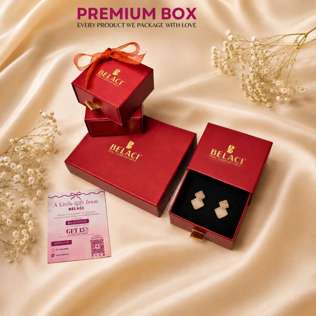 BELACI jewelry gift packaging set on a champagne satin background, featuring red boxes with gold branding, one open box displaying a pair of gold square stud earrings, and a pink gift card.