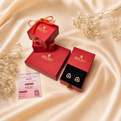 Belaci brand red gift boxes and an open box displaying gold heart-shaped earrings on a black insert, arranged on a beige satin fabric with baby's breath flowers and a gift card.