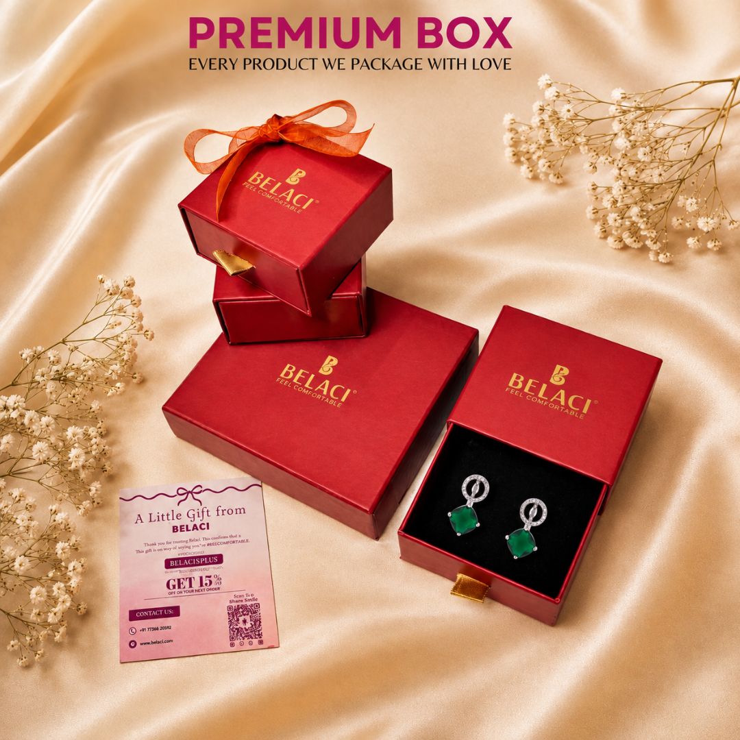 BELACI premium gift packaging set featuring red boxes, an open box displaying green gemstone earrings, and a promotional card on draped champagne satin fabric with baby's breath flowers.
