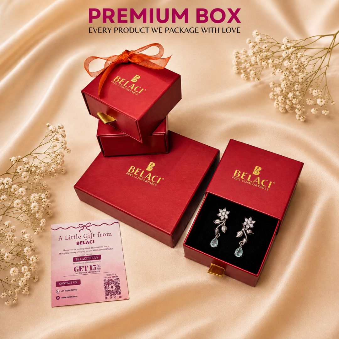 Belaci premium jewelry packaging set featuring red gift boxes, an open box displaying silver floral earrings with blue teardrop drops, a pink gift voucher, and decorative baby's breath flowers on a beige satin background.