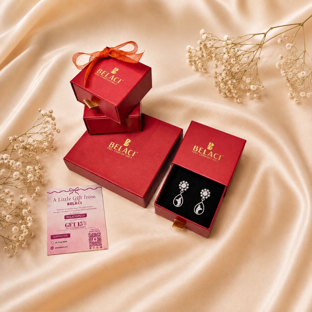 Silver teardrop flower earrings displayed in an open red Belaci gift box, alongside other closed red Belaci boxes and a pink gift card, arranged on a beige satin fabric background with dried baby's breath flowers.