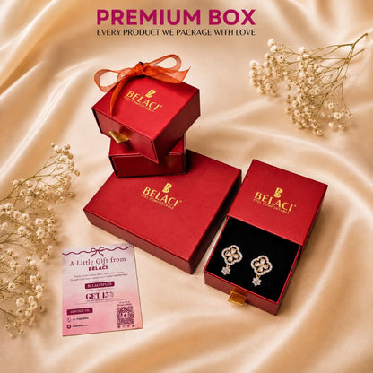 Red Belaci premium jewelry gift boxes arranged on a champagne satin background with dried baby's breath flowers. One open box displays silver floral stud earrings on a black insert, and a pink promotional card is placed nearby.