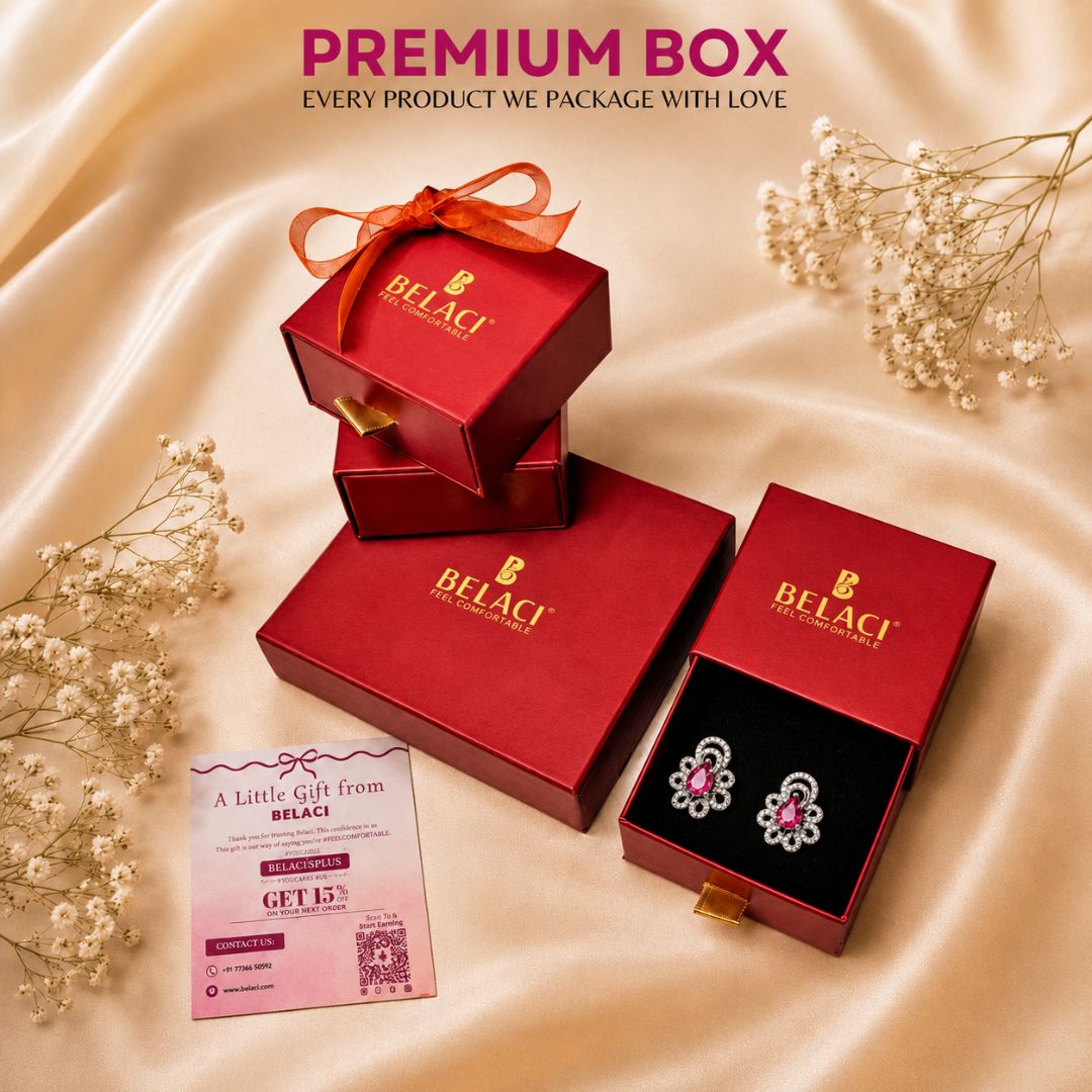 BELACI premium jewelry gift packaging set on draped champagne satin fabric with baby's breath flowers. The set includes a red gift box tied with an orange ribbon, a red gift box with a gold clasp, an open red box displaying silver and pink floral stud earrings on a black insert, and a pink promotional card.