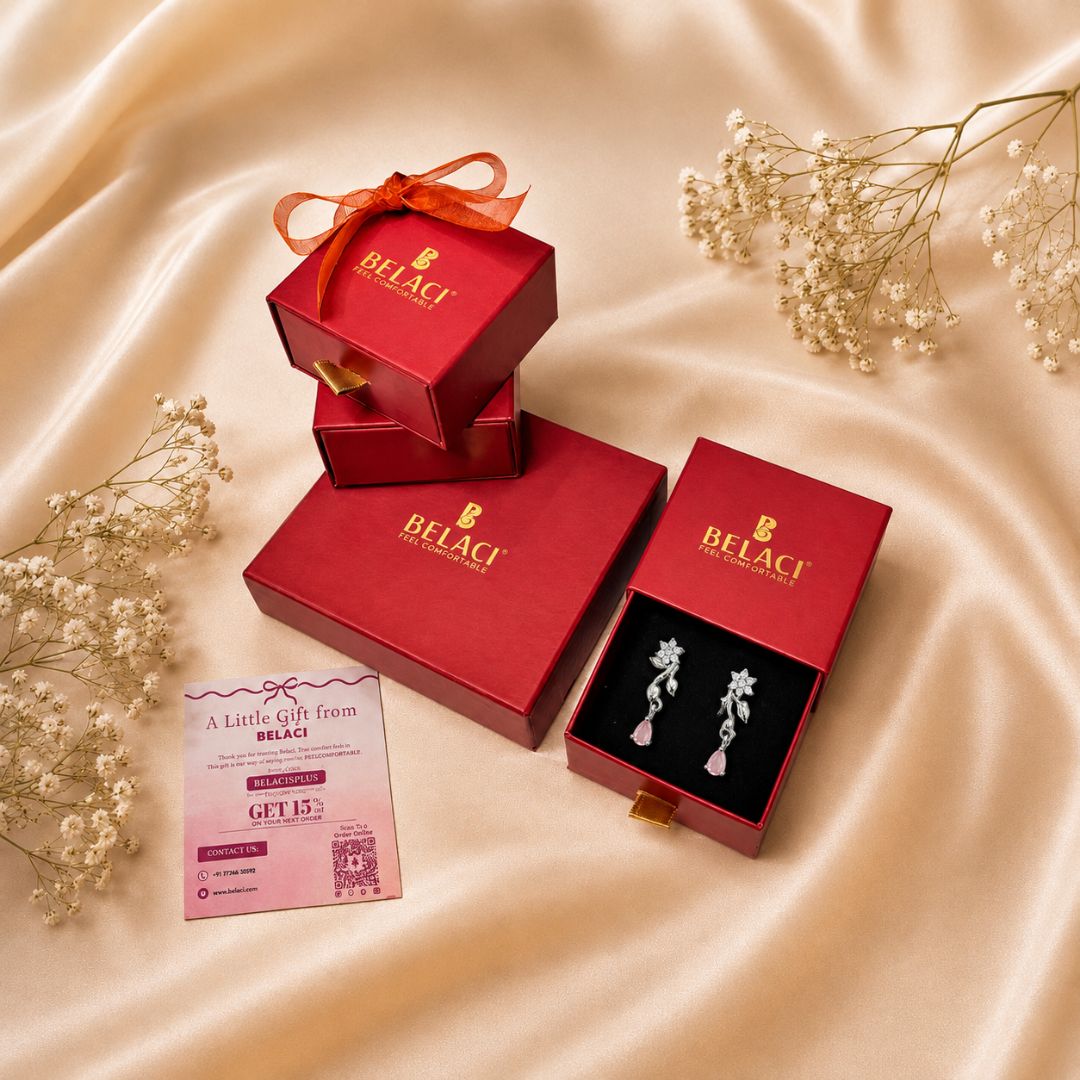 A pair of silver-toned floral earrings with pink pearl-like drops, displayed in an open red BELACI jewelry box on a beige satin fabric background. Additional red BELACI gift boxes, a pink gift card, and dried baby's breath flowers are arranged around them.