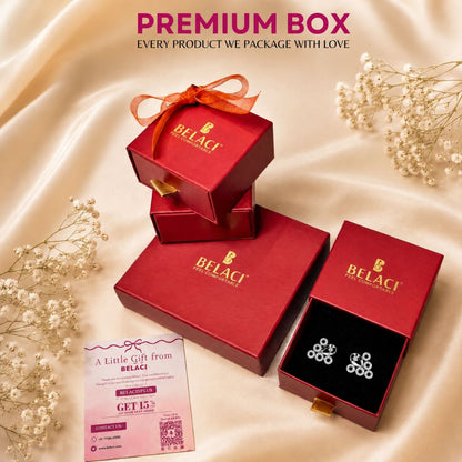BELACI jewelry gift packaging set on a champagne satin background with baby's breath flowers. The set includes a stack of red gift boxes with an orange ribbon, an open red box showing silver stud earrings on a black insert, a pink gift card, and the text 'PREMIUM BOX EVERY PRODUCT WE PACKAGE WITH LOVE' at the top.
