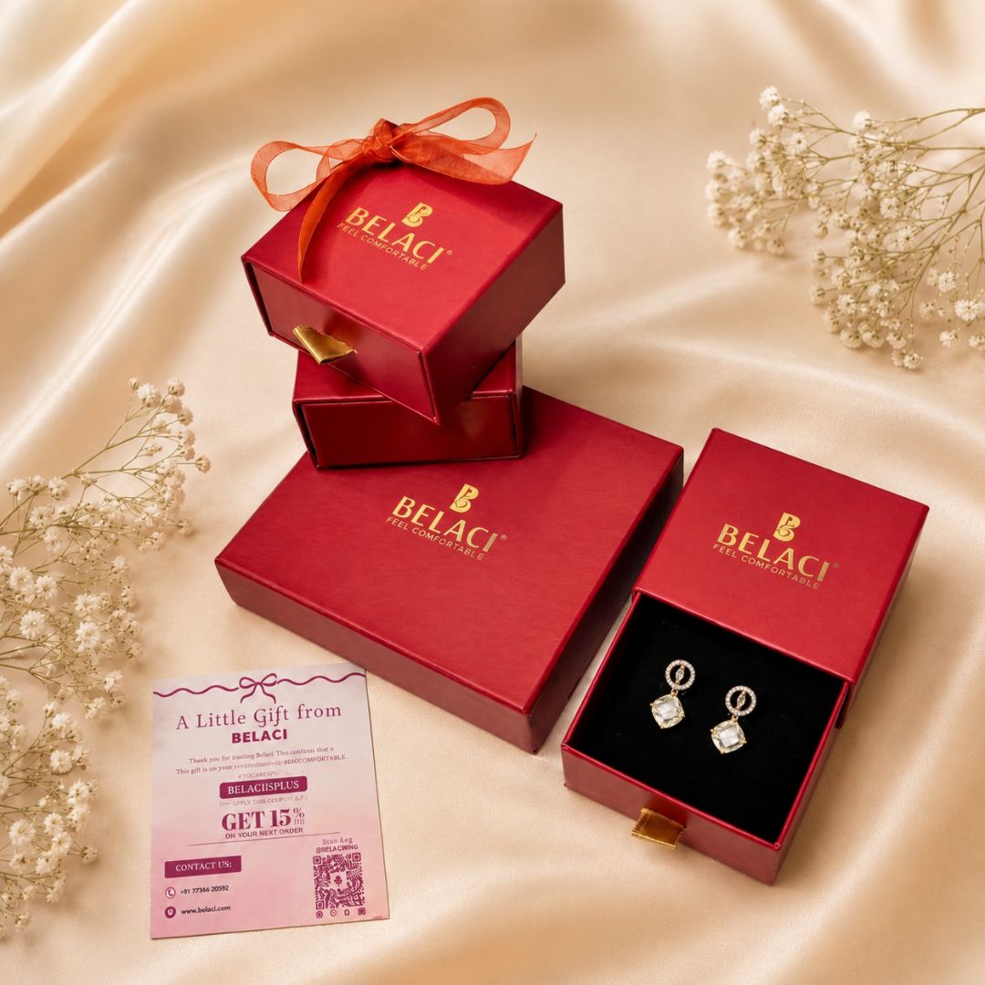 BELACI brand jewelry gift set featuring a pair of silver hoop earrings with clear gemstones in a red presentation box, alongside stacked and open red gift boxes with BELACI branding and a pink promotional card on a beige satin background.