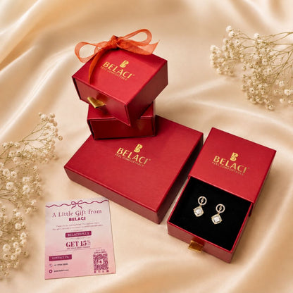 BELACI brand jewelry gift set featuring a pair of silver hoop earrings with clear gemstones in a red presentation box, alongside stacked and open red gift boxes with BELACI branding and a pink promotional card on a beige satin background.